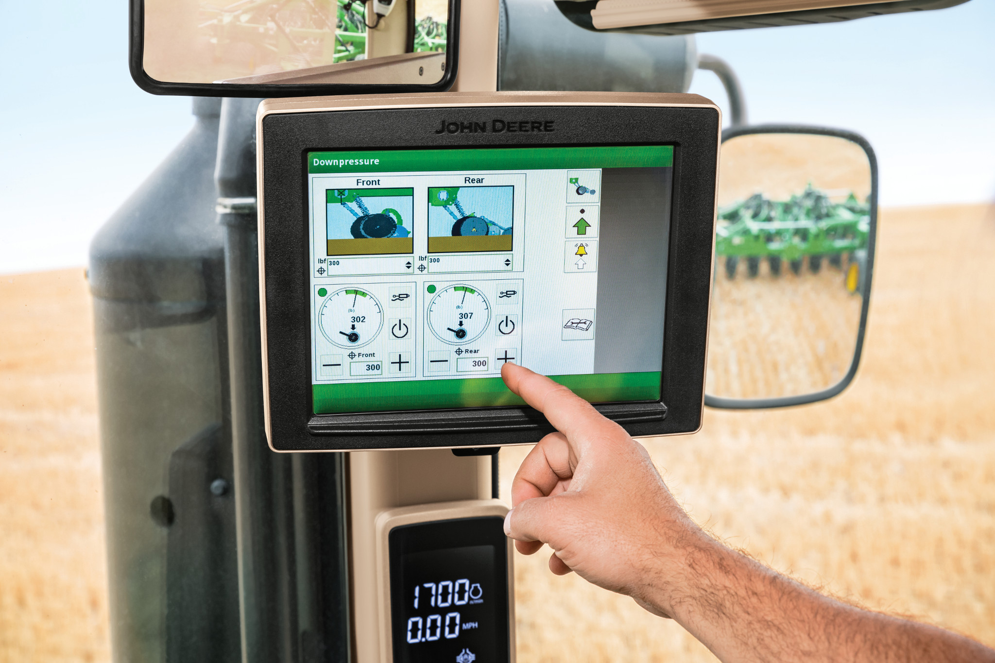 Close-up of a John Deere tractor touchscreen displaying downforce controls with gauges and diagrams as a hand presses the screen, with a field and green farm equipment visible through the cab windows and mirrors