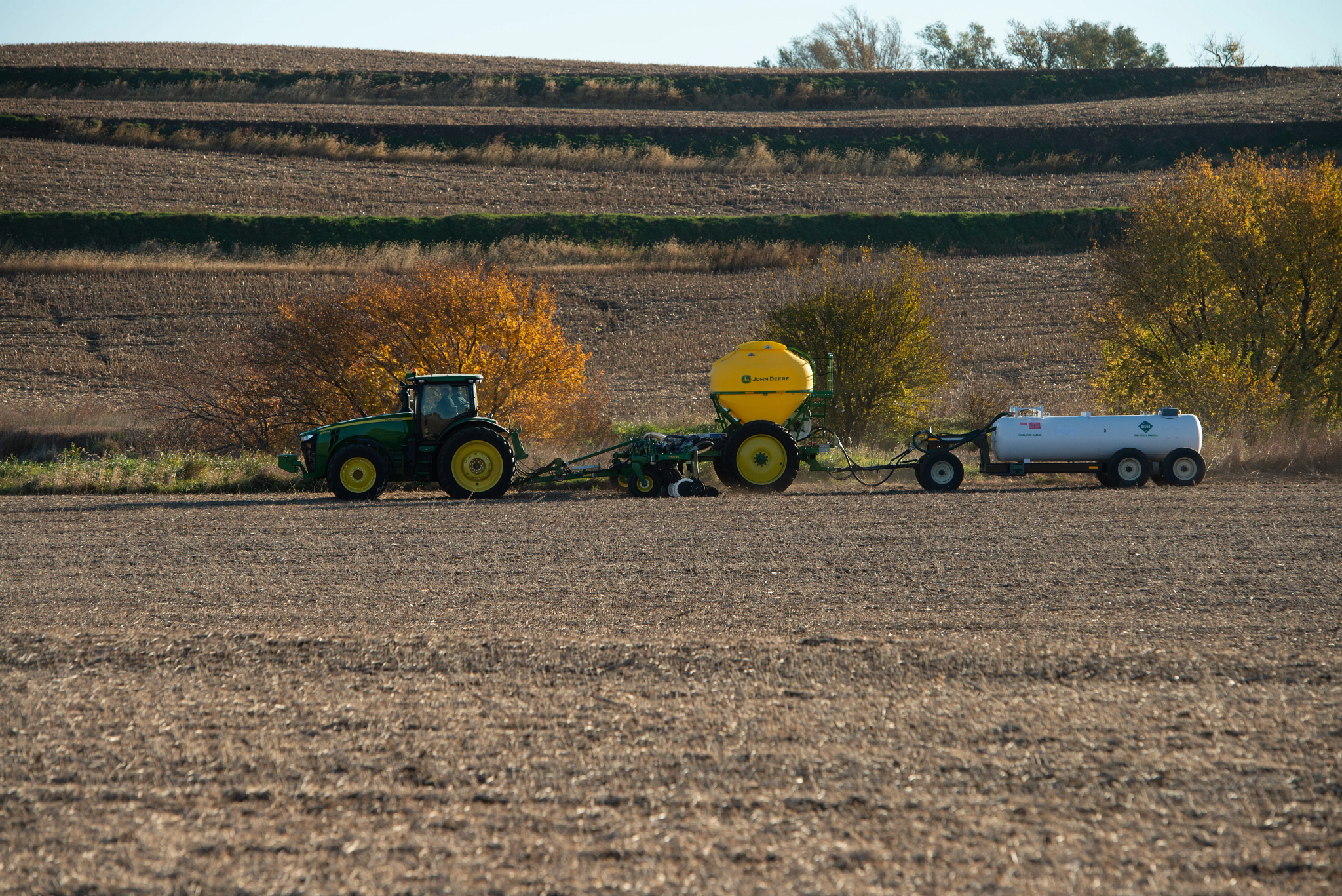 John Deere tractor pulling a nutrient applicator and an anhydrous ammonia tank across a harvested field, with terraced hills and autumn trees in the background