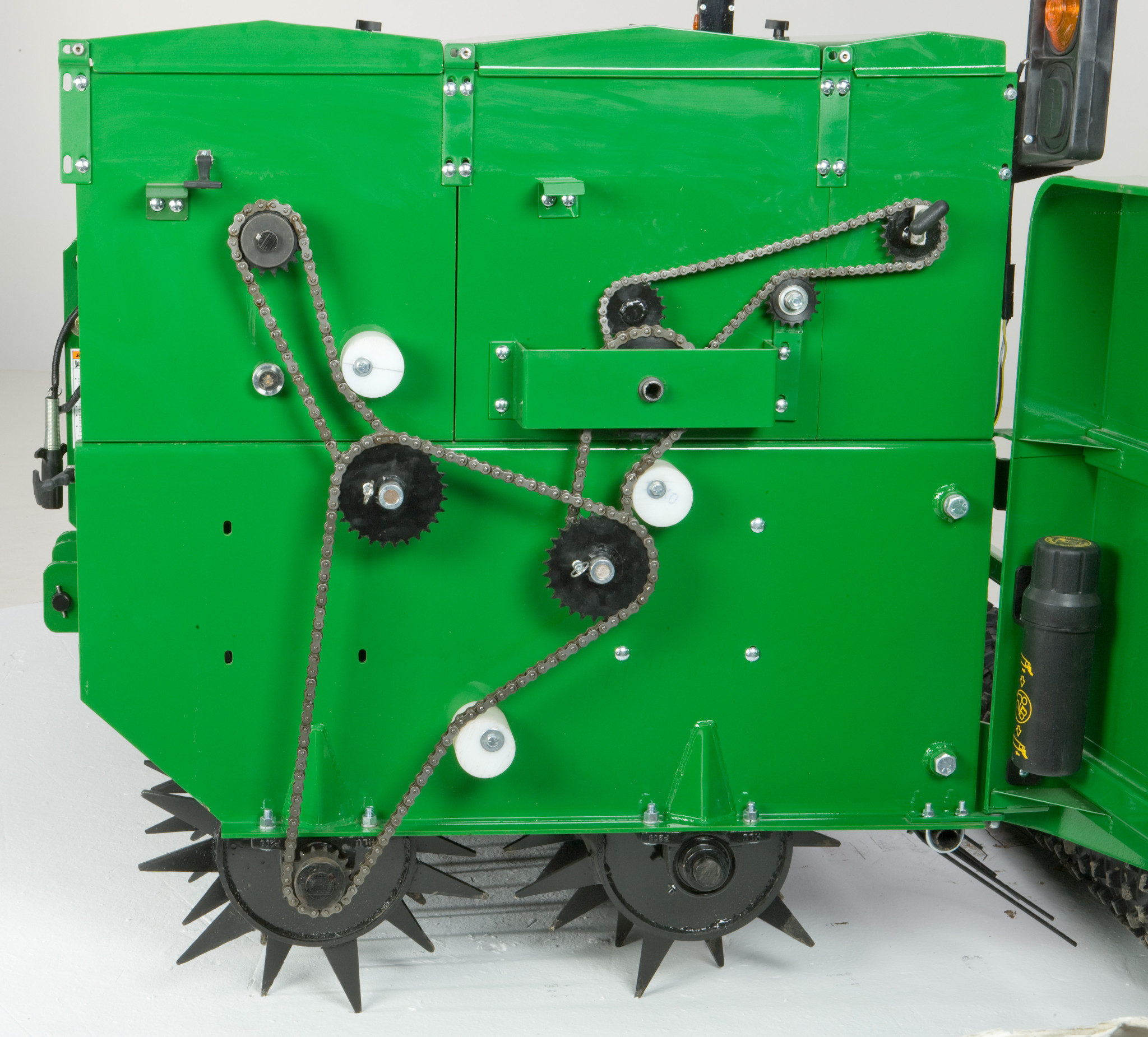 Side view of a green seeding machine showing an exposed chain‑and‑sprocket drive system with gears, idler wheels, and tensioners, above spiked ground‑engaging wheels