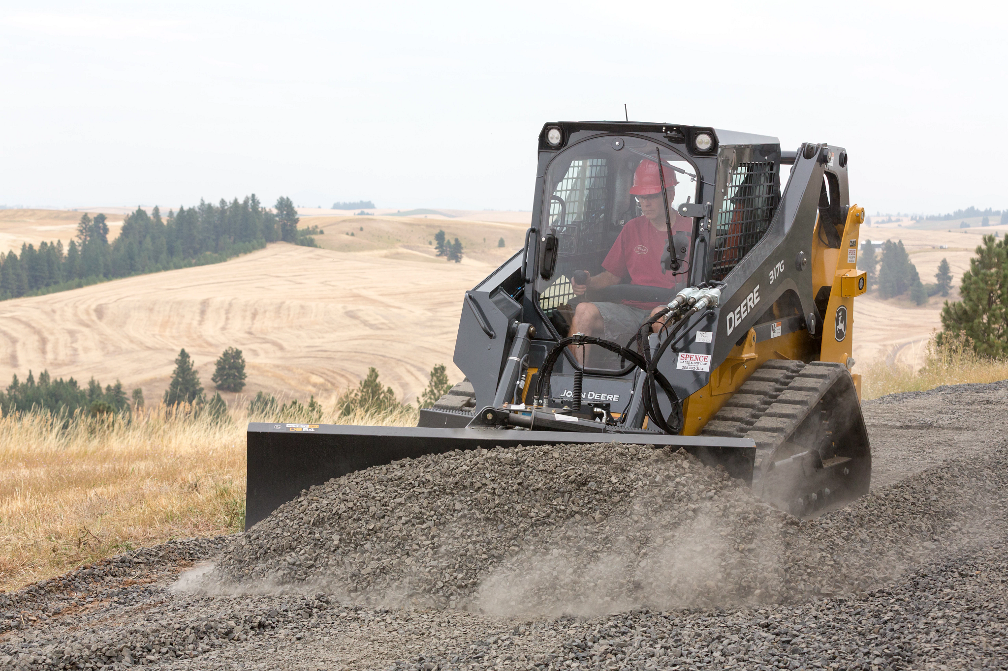 Tracked skid steer loader with a wide front blade pushing a pile of gravel on a sloped landscape, operated from an enclosed cab.
